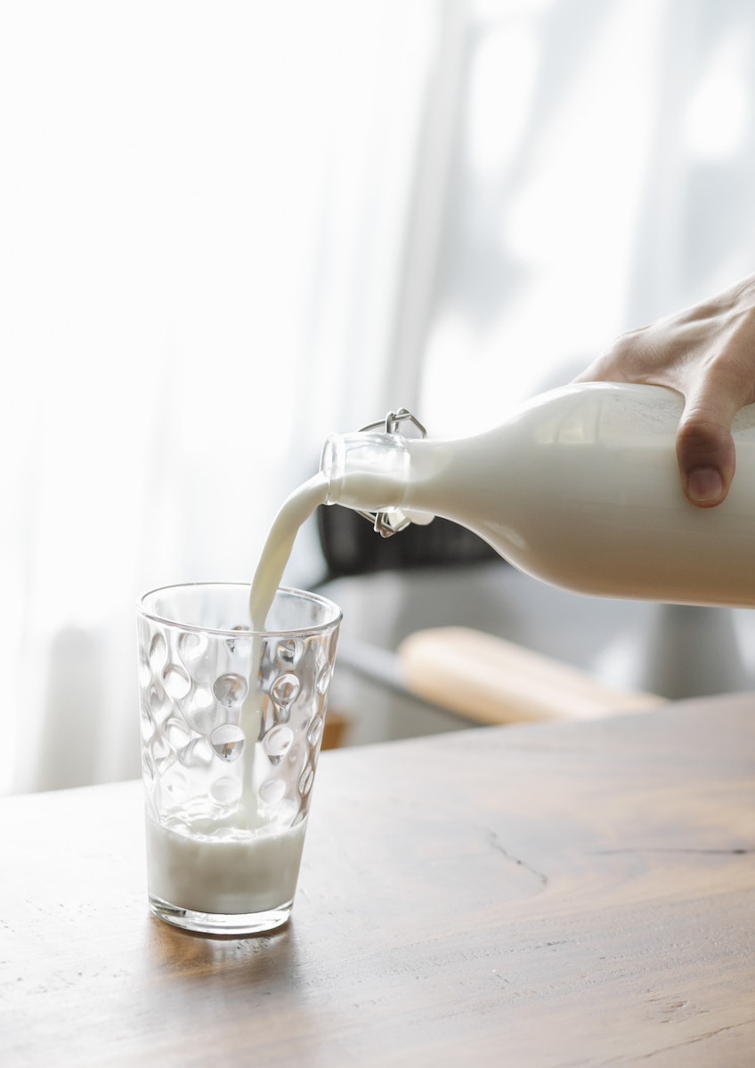 Photo by Charlotte May Crop faceless person pouring fresh cold milk from bottle into transparent clean glass placed on wooden table